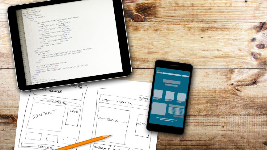 Auf einem Holztisch liegt ein Blatt Papier mit einer aufgezeichneten Website-Skizze und ein Tablet mit Programmierungscode.