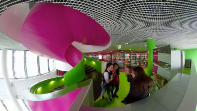 Vier Studentinnen stehen unterhalten sich neben einer Wendeltreppe in der Universitätsbibliothek
