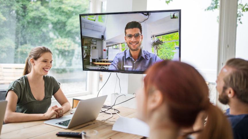 Hochschulmitarbeitende sitzen in einem Meetingraum und sehen auf dem Bildschrim einer Online-Präsentation zu.